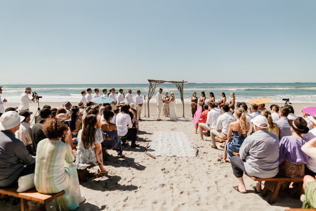 Wedding on a new zealand beach with lots of guests sitting on bench seats